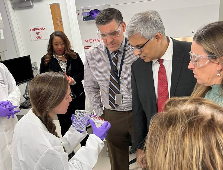 Group of people in a laboratory setting view a well plate held by a scientist in a lab coat and latex gloves.