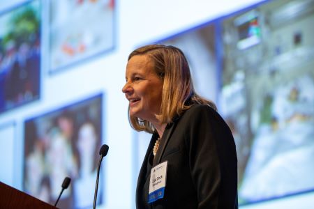 Lisa Deck speaks at lectern about her experience with a rare disease at Rare Disease Day at NIH 2020. Images related to her diagnostic journey are projected on a large screen behind her.