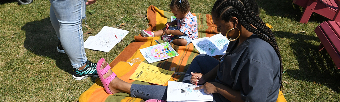 A family sits on a blanket coloring pages from the “Sofia Learns About Research” activity book. 