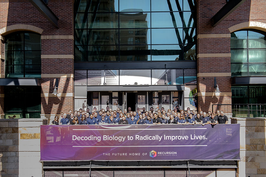 The Recursion team poses outside its new headquarters with a banner reading "Decoding Biology to Radically Improve Lives."