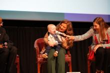 Baby KJ sits on Nicole Muldoon’s lap while Dr. Rebecca Ahrens-Nicklas hold a microphone up to them.