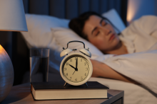 Close-up of a bedside table with an analog clock, a glass of water, and a book, while a person lies asleep in bed.